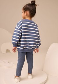 Gestreifter blauer Sweatshirt mit Ballonärmeln und geripptem Saum, kombiniert mit strukturierten blauen Leggings und weißen Sneakern. Neutraler Hintergrund.