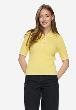 Junge Frau mit langen Haaren, die lächelt, trägt ein gelbes Polohemd mit kurzen Ärmeln und schwarze Hose, steht mit den Händen hinter dem Rücken.