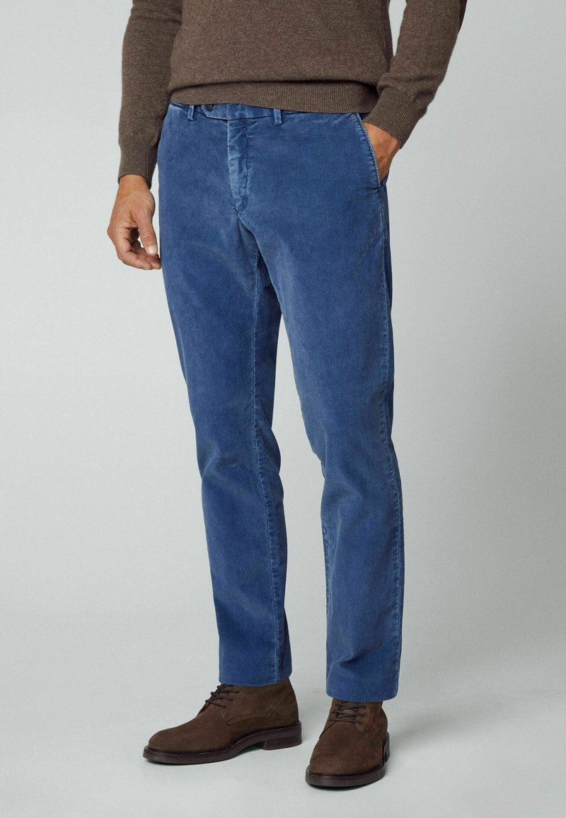 Homme portant un pantalon en velours côtelé bleu, un pull marron et des bottines lacées en suède marron, debout avec une main dans la poche sur un fond gris.