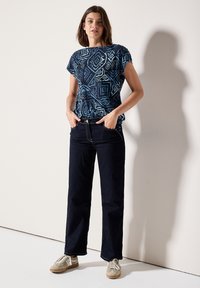 Top s geometrickým vzorem v námořnické modři a krátkým rukávem, spárovaný se tmavými džíny a světlými teniskami. Modelka stojí s rukama v kapsách.