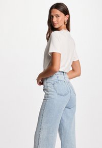 Mujer con cabello largo castaño, usando pendientes de oro, una blusa blanca de manga corta y unos vaqueros de tiro alto de denim azul claro, de pie frente a un fondo blanco.