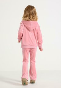 Růžová velurová mikina s kapucí, spárovaná s odpovídajícími zvonovými kalhotami. Látka je měkká s texturovaným vzhledem. Celý outfit doplňují volnočasové boty.