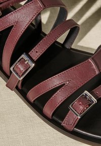 Burgunderfarvede læderstrop sandaler med en mørk sål, der har metalspænder og strukturerede stropper arrangeret i et zigzag-mønster.