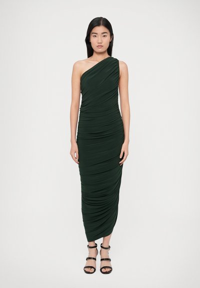 Robe longue longueur au sol, vert foncé, à une épaule, avec une texture froncée. Accessoirisée avec des sandales noires à bride cheville.