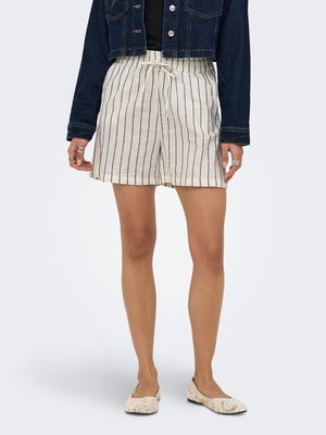 Kvinde iført hvide shorts med sorte lodrette striber, bundet talje, en mørk denimjakke og cremefarvede hæklede flade sko stående på en ensfarvet baggrund.