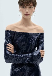 Marinblå off-shoulder klänning med paljetter och långa ärmar, som har en figurnära silhuett och utsvängda ärmslut. Slät textur med reflekterande paljetter.