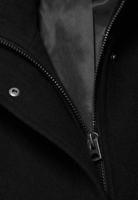 Gros plan d'une fermeture éclair noire de veste partiellement fermée, montrant une doublure en tissu lisse et un bouton-pression en métal sur la couche extérieure en laine.