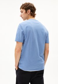 Junger Mann mit kurzen braunen Haaren, der ein hellblau gestreiftes T-Shirt und schwarze Hosen trägt, mit Händen in den Taschen steht und zur Seite schaut.