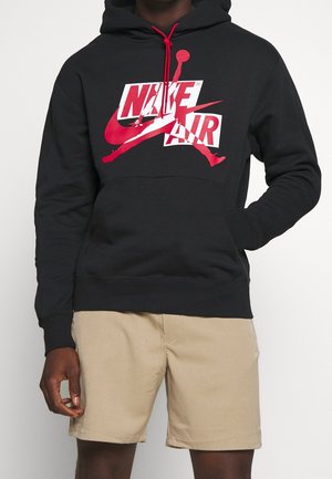 Schwarzer Kapuzenpullover mit rotem Nike Air-Logo, roten Kordeln, kombiniert mit beigen Shorts, getragen von einer Person mit einer Hand in der Tasche.