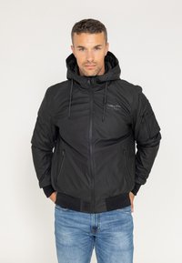 Czarna ocieplana kurtka z kapturem, zamkami i elastycznymi mankietami. Posiada logo na piersi oraz podwójne kieszenie boczne, zestawiona z niebieskimi dżinsami.