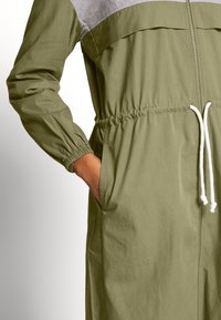 Chaqueta de cremallera verde oliva con un panel superior gris, puños elásticos, cintura con cordón y bolsillos laterales. Tejido suave y ligero.