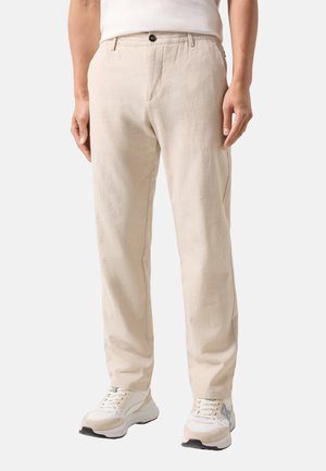 Mężczyzna w beżowych casualowych spodniach i białych trampkach, stojący na tle jednolitego jasnego tła.