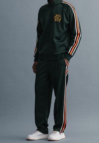Chándal verde de tejido suave, con rayas naranjas y blancas en las mangas y los pantalones, y un logo en el pecho. Zapatillas blancas.