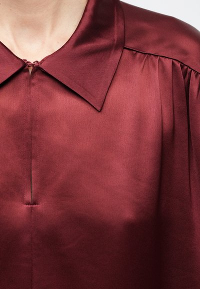 Close-up af en person iført en bordeaux rød satinbluse med bred krave, frontslids og folder ved skuldersømmen.
