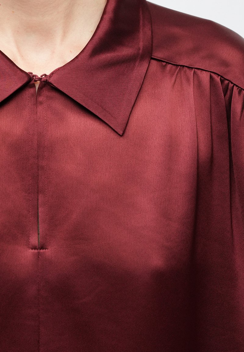 Close-up of a person wearing a burgundy satin blouse with a wide collar, front slit, and gathers on the shoulder seam.