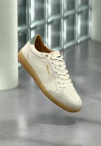 Άσπρο δερμάτινο αθλητικό παπούτσι με λαστιχένια σόλα σε χρώμα σοκολάτας, πράσινη ταμπέλα στη φτέρνα και το όνομα της μάρκας "Blauer", εμφανίζεται να αιωρείται σε εσωτερικό χώρο με φόντο γκρι πάτωμα και παράθυρο.