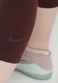 Zbliżenie na nogę w bordowych legginsach Nike i jasnoróżowych butach do biegania Nike na tle jasnej skóry.