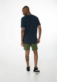 Polo shirt marine avec des accents blancs, associé à un short vert. Baskets montantes noires, tatouages visibles sur les jambes, fond neutre.