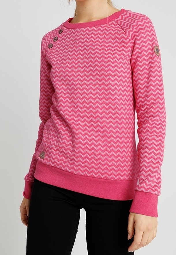 Femme portant un pull rose à motif zigzag avec des boutons sur l’épaule et un pantalon noir, debout devant un fond uni.