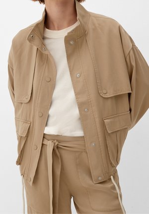 Persoon draagt een beige utility jas met grote klepzakken, wit overhemd en bijpassende beige broek, in de taille gestrikt met een stoffen riem.