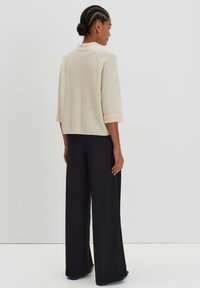 Maglione beige a costine con maniche a tre quarti, indossato con pantaloni neri larghi e comodi. Design minimalista con polsini arrotolati.