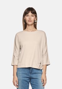Light beige long-sleeve top with a relaxed fit and a single patch pocket. Soft fabric and a round neckline enhance its simplicity.