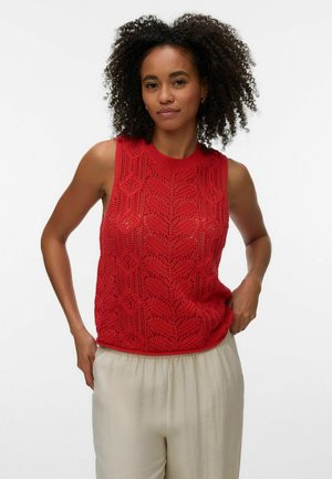 Femme aux cheveux bouclés portant un haut sans manches rouge à motif en tricot et un pantalon ample beige, debout devant un fond blanc uni.