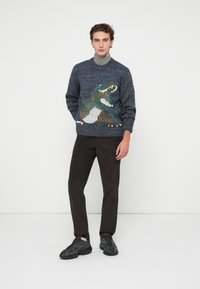 Pulover tricotat în nuanțe de albastru și verde, având un design cu dinozaur, asortat cu pantaloni negri din catifea și adidași masivi negri.