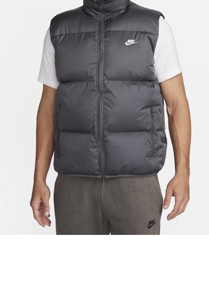 Homme portant une veste matelassée noire sans manches Nike sur un t-shirt blanc à manches courtes et un pantalon de jogging gris Nike, debout avec les bras relâchés.