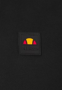 Tissu noir avec un patch carré cousu comportant un logo en demi-cercle jaune et rouge au-dessus du texte « ellesse » en noir.