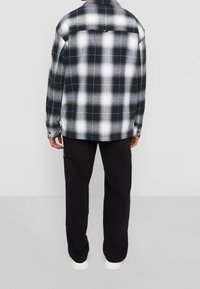 Camicia di flanella a quadri bianco e nero con chiusura frontale con bottoni visibili e dettagli sui polsini, indossata con pantaloni neri, vista da dietro.