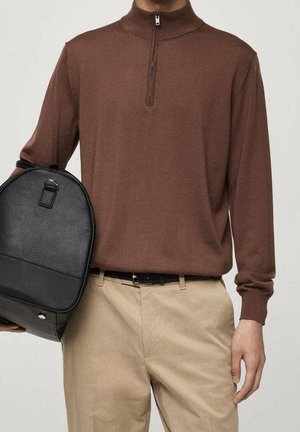 Personne portant un pull zippé à manches longues marron et un pantalon beige, tenant un sac à dos en cuir noir à ses côtés.