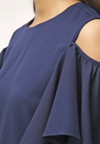 Tmavě modrý top vyrobený z texturované látky; má široké volánové rukávy a design s vykrojenými rameny. Jednoduchý kulatý výstřih.