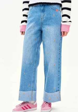 Jasnoniebieskie dżinsy o szerokich nogawkach, ozdobione małym różowym haftem kwiatowym i surowymi zakończeniami, noszone z różowymi trampkami i paskowanym swetrem z różowymi mankietami.