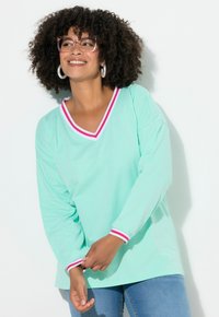 Donna con i capelli corti e ricci che indossa occhiali rosa, orecchini a cerchio, un top a maniche lunghe verde menta con un bordo a righe, e jeans blu mentre sorride.