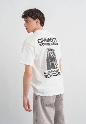 Ung mann med kort krøllete hår som har på seg en hvit Carhartt T-skjorte med svart dekk og tekstgrafikk på baksiden, stående mot en ensfarget bakgrunn.