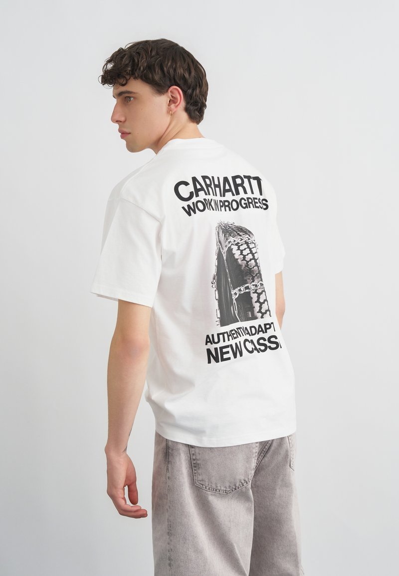 Νέος άνδρας με κοντά κατσαρά μαλλιά που φοράει λευκό T-shirt Carhartt με μαύρο γραφικό ελαστικού και κείμενο στην πλάτη, στέκεται μπροστά σε μια απλή φόντο.