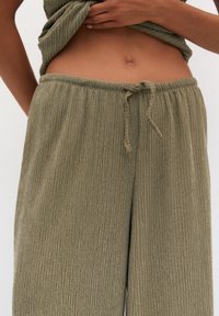Pantalon large couleur vert olive en tissu texturé à côtes. Taille élastique avec cordon de serrage pour ajustement. Poches latérales visibles.