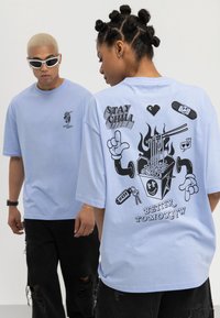 To personer iført lyseblå "Stay Chill" t-skjorter; den ene viser ryggen med en tegneserie av nudler, flammer og teksten "Stay Chill" og "Better Tomorrow."