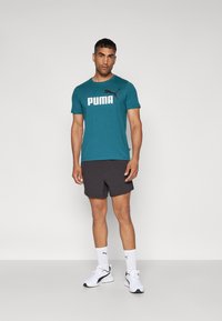Žalias trumpomis rankovėmis marškinėliai su balta "PUMA" logotipu, derinami su juodais šortais ir baltais sportiniais batais su juodais akcentais. Paprastas, modernus dizainas.