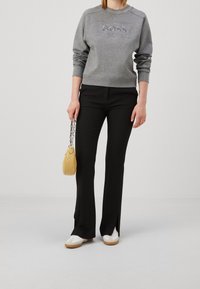 Felpa grigia con logo in rilievo, abbinata a pantaloni neri svasati. La modella tiene una borsa a tracolla gialla trapuntata con catena. Sneakers bianche.