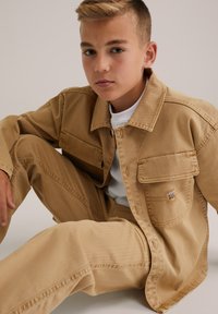 Najstnik sedi s kolenom dvignjenim, oblečen v bež denim jakno in hlače preko bele majice, nevtralen ozadje.