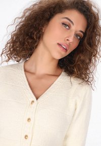 Cremfarbener Strickcardigan mit strukturiertem Muster, V-Ausschnitt und fünf Holzknöpfen an der Vorderseite. Weiches, gemütliches Aussehen.