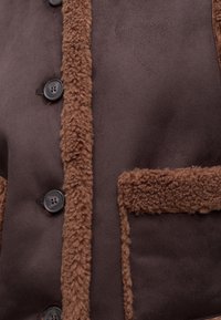 Κοντινή λήψη ενός καφέ παλτού shearling με υφή στο περίγραμμα κατά μήκος των κουμπιών και των άκρων των τσεπών.