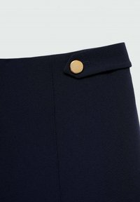 Sukně námořnické modři s hladkým texturem, s kovovou knoflíčkem zlaté barvy v pase a zúženým střihem. Detailní pohled na oblast pasu.