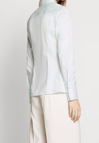 Chemise à manches longues bleu clair en satin avec un col contrasté, coupe ajustée et poignets boutonnés, mettant en valeur une texture lisse et des lignes épurées.