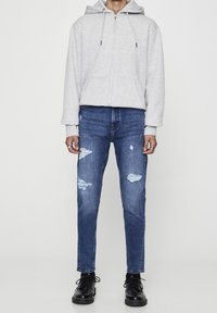 Sudadera con cremallera de color gris claro con bolsillos tipo canguro, combinada con jeans azules desgastados que tienen rotos y un corte ajustado, complementada con botas negras de cordones.
