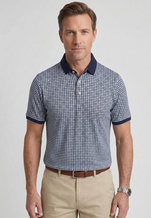 Man wearing blue and white checkered polo shirt with navy collar and cuffs, beige pants, brown belt, and silver wristwatch, standing.