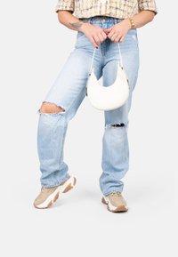 Sac à main blanc, courbé, tenu à la main, associé à un jean bleu clair déchiré avec des trous aux genoux et des baskets beiges à semelles épaisses.