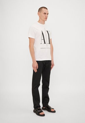 Hvid T-shirt med sort "AX" og "ARMANI EXCHANGE" grafik, parret med sorte bukser og sorte sandaler, præsenteret mod en ensfarvet baggrund.